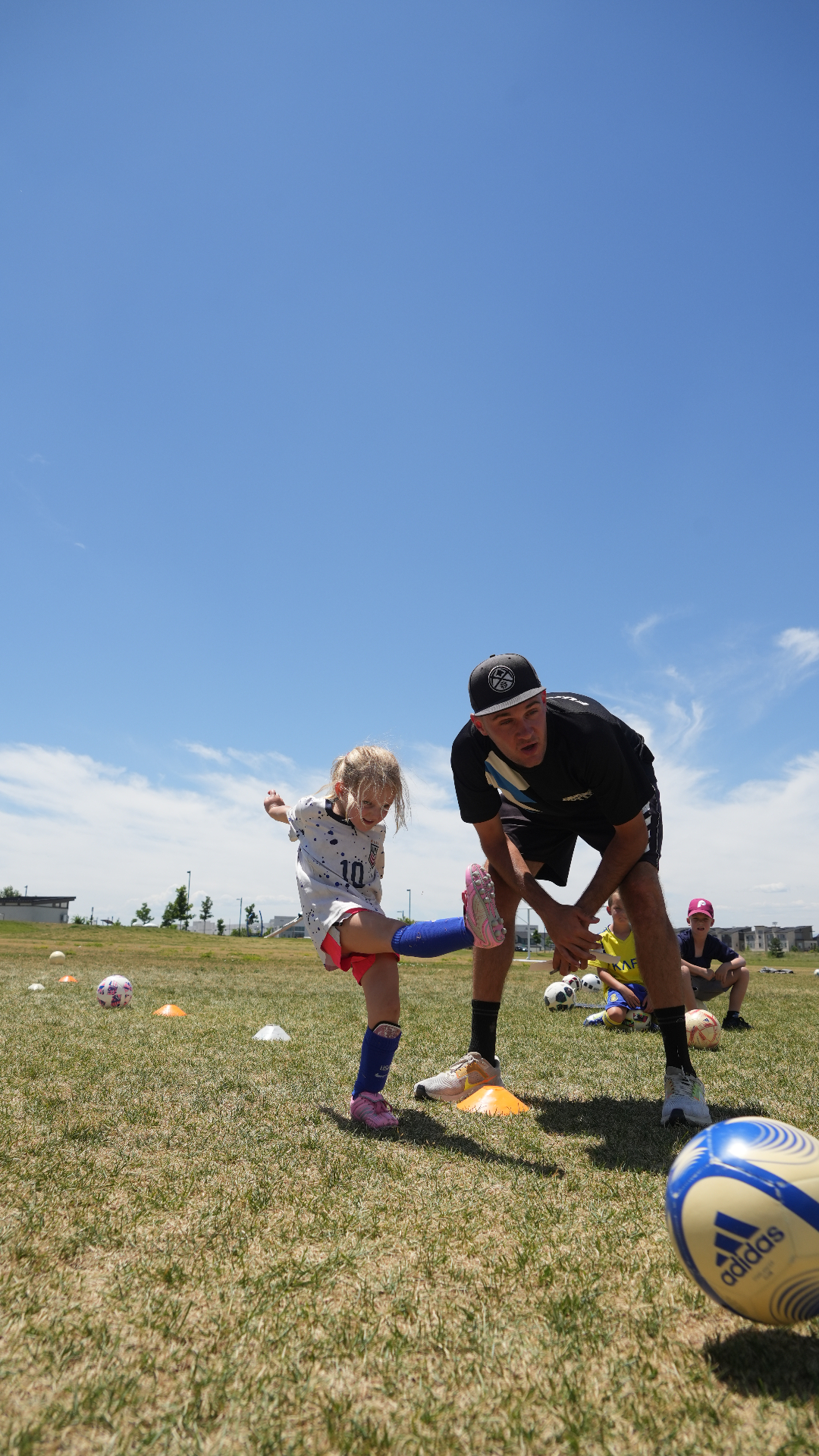 Young girl kicking a soccer ball with her coach crouched beside her